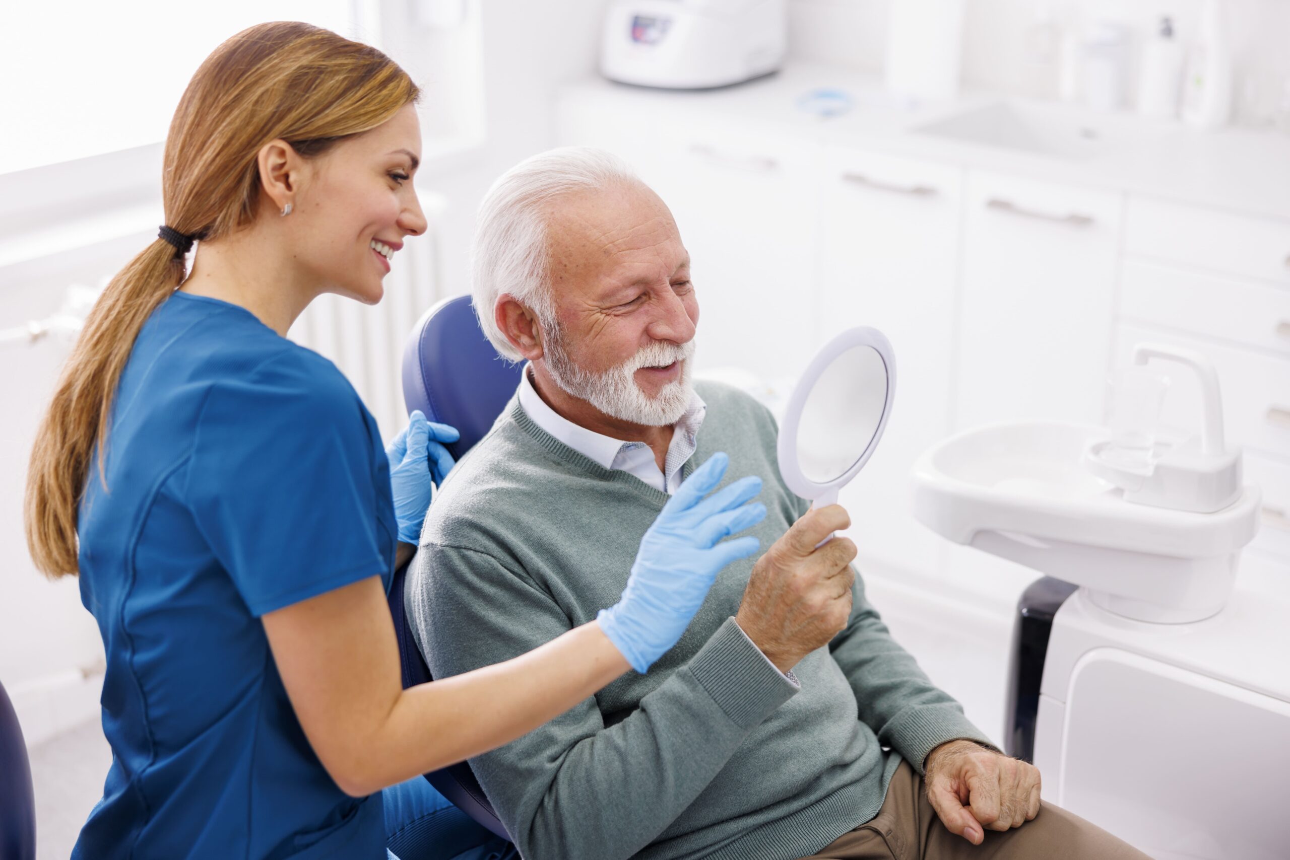 Senior man smiling into hand mirror held by dental hygienist